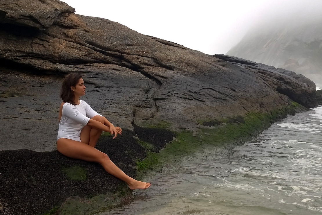 8 motivos e a Alezzia sentada na pedra em frente ao mar em um dia nublado